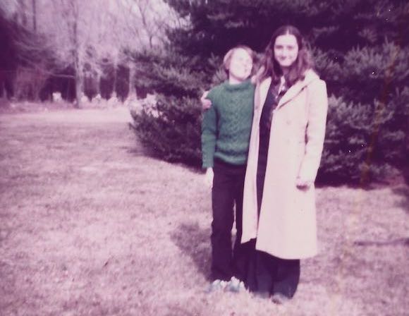 Young Peter and his teenage sister outdoors on a lawn with a pine tree behind. Peter's sister has her arm around his shoulder. Photo is from the mid 1970s.