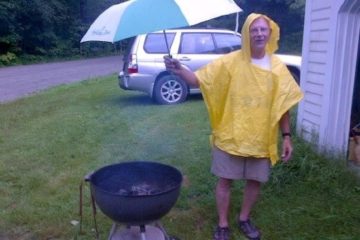 man standing in a rainstorm wearing a yellow slicker and holding a big umbrella over a charcoal barbecue