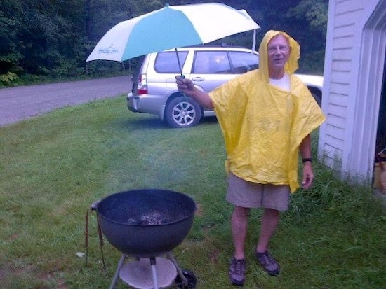 man standing in a rainstorm wearing a yellow slicker and holding a big umbrella over a charcoal barbecue