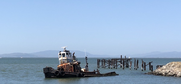 A dilapidated tugboat rests in the shallows a few yards from shore beside the ruins of a long-decayed pier.