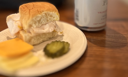 A small turkey sandwich next to some cheese slices and pickle slices on a plate, with a can of beer behind it