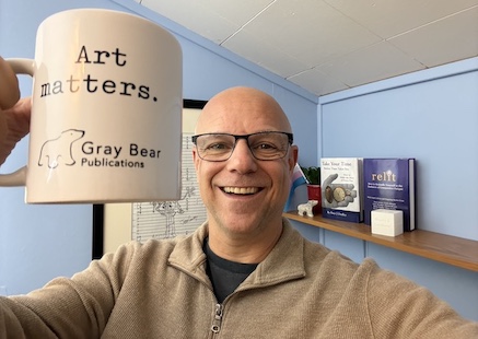 Peter smiling and holding up a Gray Bear Publications branded mug saying "Art matters." with a few of his other books on the shelf behind him in his office. He will not be reduced!
