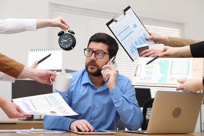 A man sits at a desk multitasking on the pone with seven colleagues each giving him a different task to do.