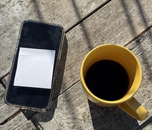 Cell phone with a blank white post-it on the screen, next to a yellow coffee mug with coffee, outdoors on a wooden table