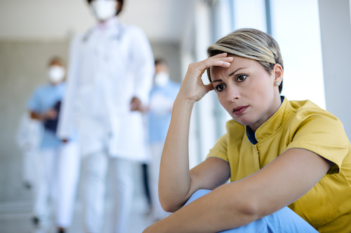 A worried woman sits on the floor of a hospital hallway as medical providers walk past, blurred in the background.