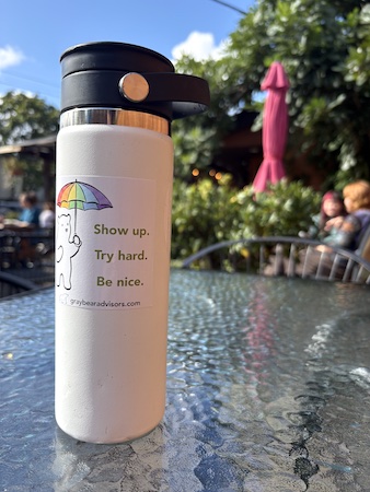 My white hot cup with a Gray Bear sticker on it and the cafe in the background. The sticker says "show up, try hard, be nice" and has Gray Bear holding a rainbow umbrella.