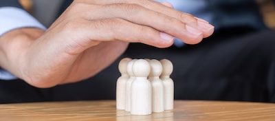 A hand hovering protectively over a number of wooden figures which represent the empathetic leadership of people being sheltered.